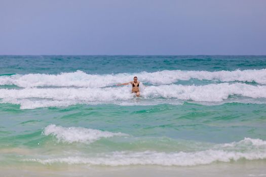 Young beautiful girl enjoys the sea and the waves, laughing raising her hands up in a black fashionable swimsuit.