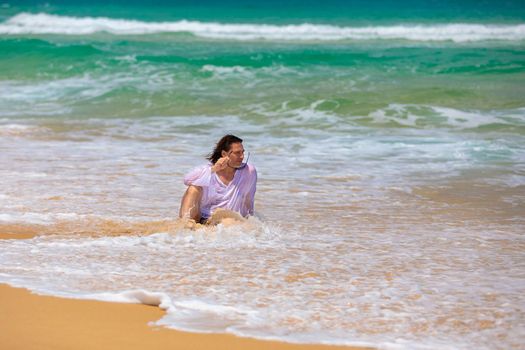 A young man with long black hair sits on the beach in the water and looks at the small waves