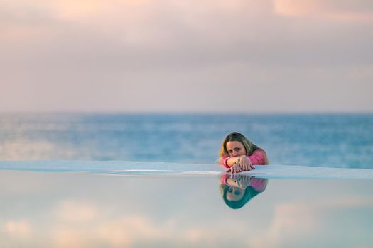 A young girl in a red shirt, at dawn by the sea pool, calm and relaxed looking at the reflection in the water. Pink clouds in the background.