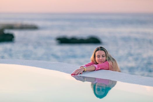 A young girl in a red shirt, at dawn by the sea pool, calm and relaxed looking at the reflection in the water. Pink clouds in the background.