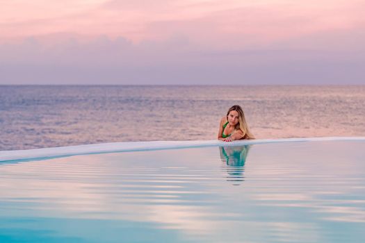 A young girl in a green swimsuit, at dawn by the sea pool, calmly and relaxed looking at her reflection in the water. Pink clouds in the background.