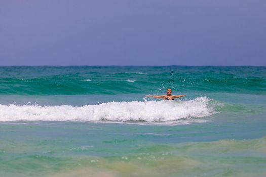 Young beautiful girl enjoys the sea and the waves, laughing raising her hands up in a black fashionable swimsuit.