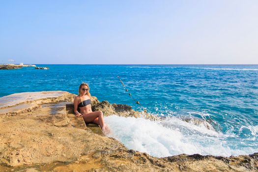 A young girl in a bathing suit and glasses sits on the steps of a cliff by the sea. Powerful waves wash over her feet. The girl laughs