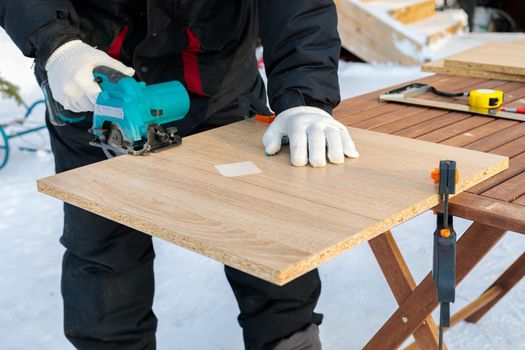 A male worker cuts a wooden panel attached to a workbench with clamps using a circular disc on a battery.