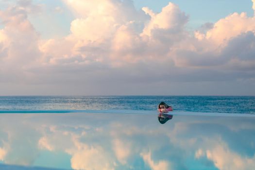 A young girl in a red shirt, at dawn by the sea pool, calm and relaxed looking at the reflection in the water. Pink clouds in the background.