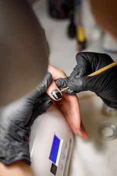 In a beauty salon, the master does a manicure, applies a pattern to the nail with a brush, black varnish with a golden pattern.