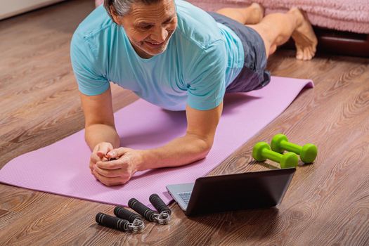 50-year-old man performs exercises while lying on the rug at home, looking at the computer. During a pandemic, a person trains in an apartment via the Internet.