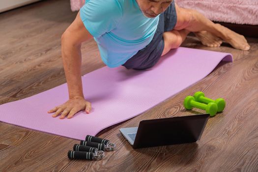 50-year-old man performs exercises while lying on the rug at home, looking at the computer. During a pandemic, a person trains in an apartment via the Internet.