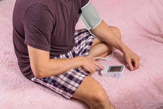 A 50-year-old man measures his blood pressure with a blood pressure monitor at home, sitting on his bed in his home clothes. Surprised by the instrument readings