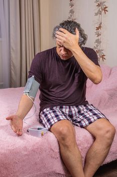 A 50-year-old man measures his blood pressure with a blood pressure monitor at home, sitting on his bed in his home clothes. Surprised by the instrument readings