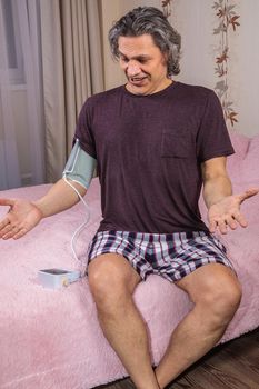 A 50-year-old man measures his blood pressure with a blood pressure monitor at home, sitting on his bed in his home clothes. Surprised by the instrument readings