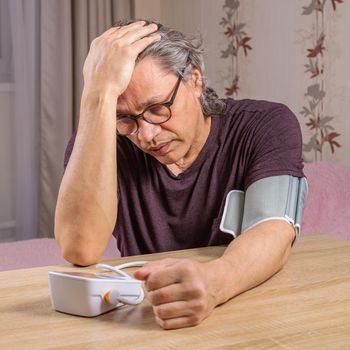 A 50-year-old man measures his blood pressure with a blood pressure monitor at home, sitting on his bed in his home clothes. Surprised by the instrument readings