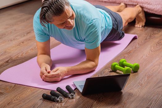 50-year-old man performs exercises while lying on the rug at home, looking at the computer. During a pandemic, a person trains in an apartment via the Internet.