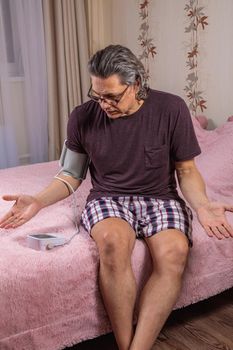 A 50-year-old man measures his blood pressure with a blood pressure monitor at home, sitting on his bed in his home clothes. Surprised by the instrument readings