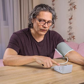 A 50-year-old man measures his blood pressure with a blood pressure monitor at home, sitting on his bed in his home clothes. Surprised by the instrument readings