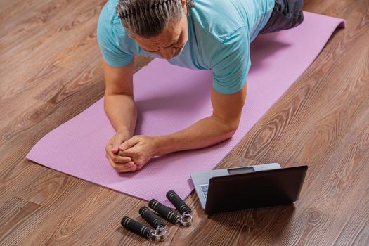 50-year-old man performs exercises while lying on the rug at home, looking at the computer. During a pandemic, a person trains in an apartment via the Internet.