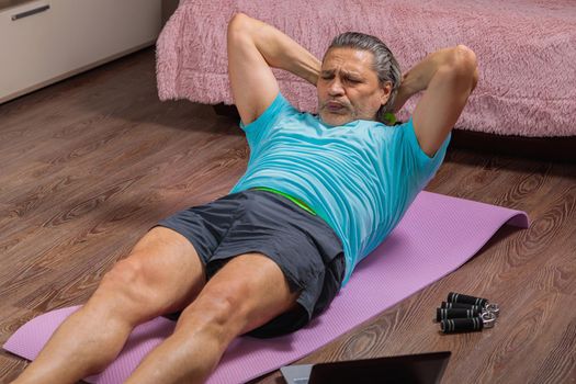 50-year-old man performs exercises while lying on the rug at home, looking at the computer. During a pandemic, a person trains in an apartment via the Internet.