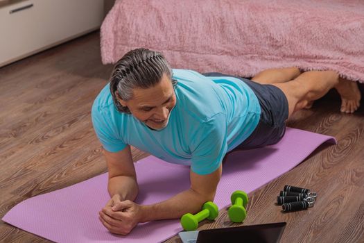 50-year-old man performs exercises while lying on the rug at home, looking at the computer. During a pandemic, a person trains in an apartment via the Internet.