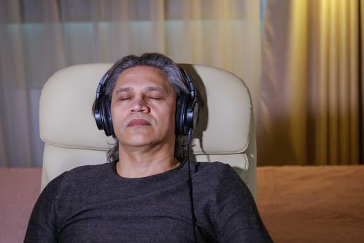 50-year-old man listens to music on headphones at home, sitting in a chair. Relaxing delight.