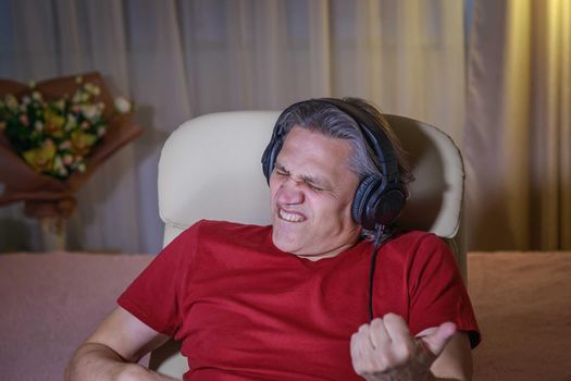 50-year-old man listens to music on headphones at home, sitting in a chair. Relaxing delight.