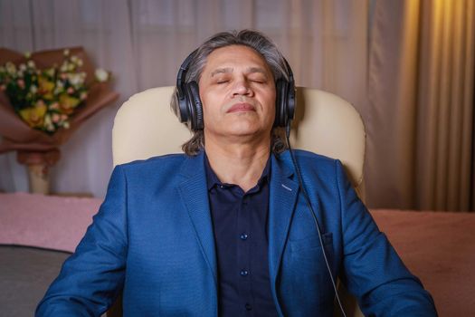 50-year-old man listens to music on headphones at home, sitting in a chair. Relaxing delight.