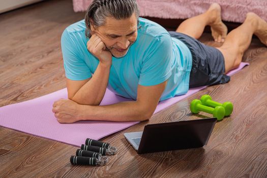 50-year-old man performs exercises while lying on the rug at home, looking at the computer. During a pandemic, a person trains in an apartment via the Internet.