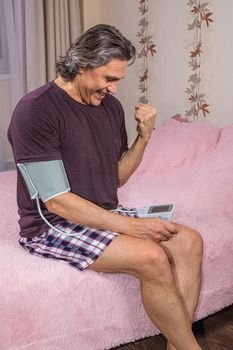 A 50-year-old man measures his blood pressure with a blood pressure monitor at home, sitting on his bed in his home clothes. Surprised by the instrument readings