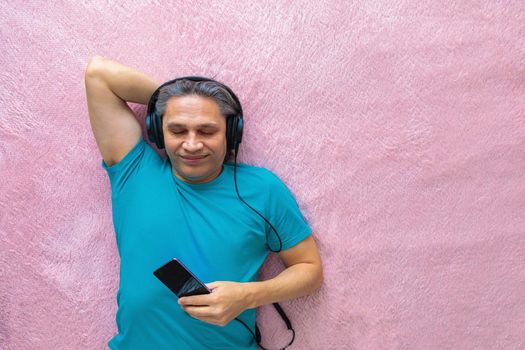 50-year-old man listens to music with headphones at home, lying on the bed. Relaxing, enjoying.