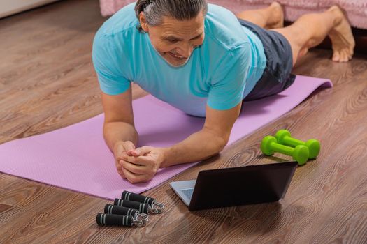 50-year-old man performs exercises while lying on the rug at home, looking at the computer. During a pandemic, a person trains in an apartment via the Internet.