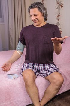 A 50-year-old man measures his blood pressure with a blood pressure monitor at home, sitting on his bed in his home clothes. Surprised by the instrument readings