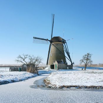 windmill in snow landscape of kinderdijk in holland under blue sky with trees and frozen canal