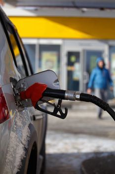 silver metallic color car refueling on winter day gas station - close-up with selective focus and blurry man silhouette in the background.