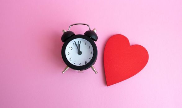 Black vintage alarm clock and red wooden heart lying on pink background.