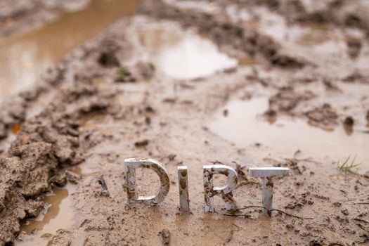 the word dirt composed of silver metal letters on wet clay surface with selective focus, background blur and linear perspective in cloudy autumn day light