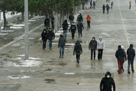 People in warm clothes and covid-19 masks walk at the waterfront with snow falling.