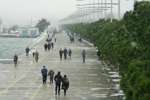 People in warm clothes and covid-19 masks walk at the waterfront with snow falling.