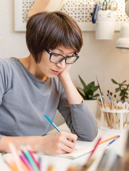 Woman with short hair cut is drawing in notebook. Calming hobby, anti stress leisure. Artist at work. Cozy workplace.