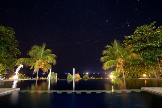 Luxury hotel with a swimming pool and palm trees under the starry sky. Long exposure, sky with stars, pool, sea, palm trees.