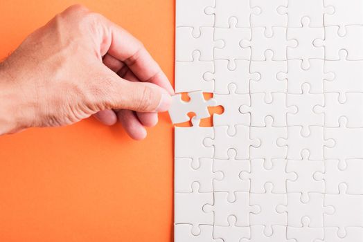 Top view flat lay of hand-holding last piece white paper jigsaw puzzle game last pieces put to place for solve problem complete mission, studio shot on an orange background, quiz calculation concept