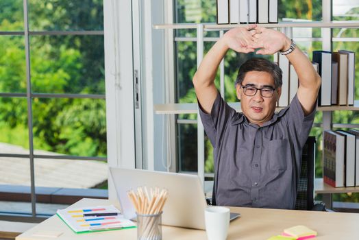 Asian businessman tired overworked he stretch oneself on the desk. senior man with eyeglasses break stretching his arms on table at his working place