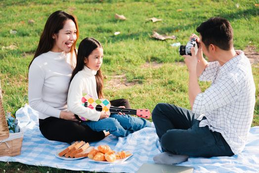 Happy Asian young family father, mother and child having fun and enjoying outdoor together sitting on the grass party with shooting photos by retro camera a picnic in the garden park on a sunny day