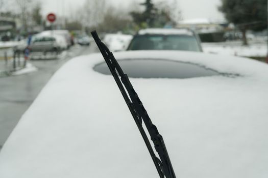 Day view of frozen windshield blade raised up against blurred background of a parked vehicle on a cold winter day.