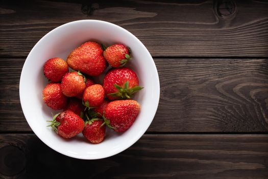 Natural ripe strawberries in a plain white bowl on a dark wooden background. Top view, copyspace.