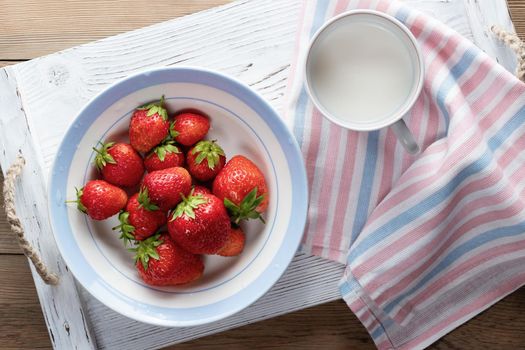 Natural ripe strawberries in a white bowl and milk in a mug on a white wooden tray. Top view.