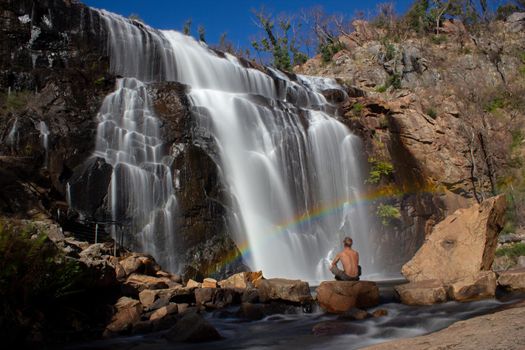 Mann, der auf einem Felsen unter einem Regenbogen vor Wasserfall, Mackenzie Falls, die Grampians, Australien sitzt