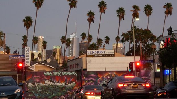 LOS ANGELES, CALIFORNIA, USA - 30 OCT 2019: Urban skyline and palms. LA city night aesthetic, graffiti painting on Vermont street. Highrise skyscrapers in downtown of metropolis. Road intersection.
