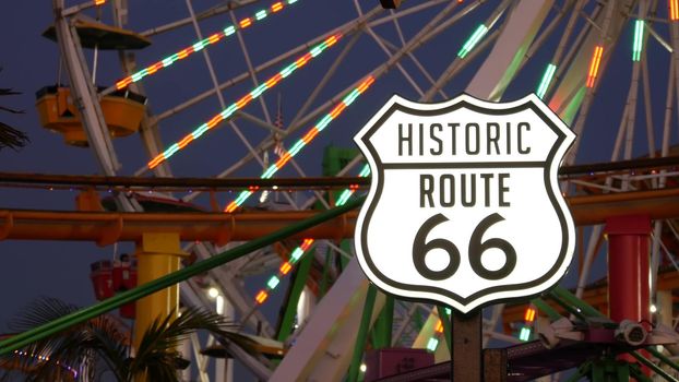 SANTA MONICA, LOS ANGELES, USA - 28 OCT 2019: Iconic road sign glowing, historic route 66. Famous california symbol, pier of pacific ocean resort. Illuminated festive ferris wheel in amusement park.