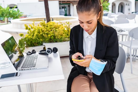 New normal freelance mobile works allows you to work anywhere with an internet connection. Modern millennial hipster woman using smartphone and laptop at bar table in the city wearing protective mask
