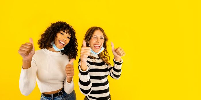 Two mixed race girls winking with thumbs up and blue medical mask thinking positive against Coronavirus pandemic isolated on yellow background copy space. Smiling confident millennial women friends