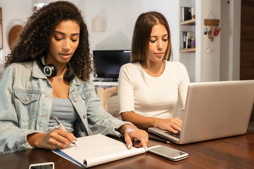 Two young millennials women self entrepreneur working from home with laptop and smartphone in the living room writing notes. New Normal internet jobs allow you to work anywhere with mobile connections